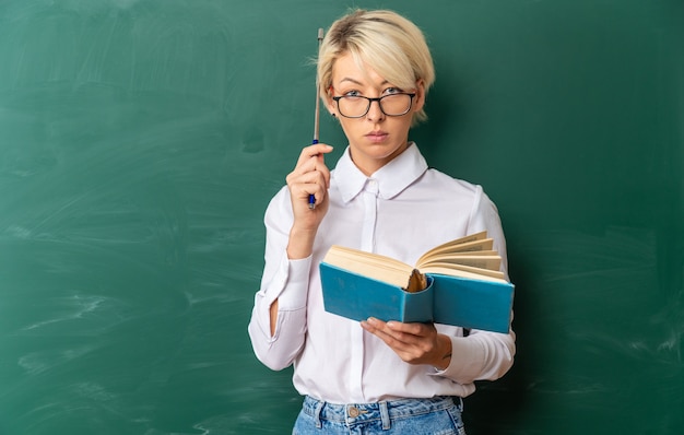 Jeune enseignante blonde sérieuse portant des lunettes en classe debout devant un tableau tenant un livre touchant la tête avec un bâton de pointeur regardant à l'avant avec un espace de copie