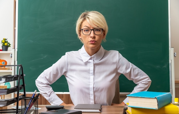 Jeune enseignante blonde stricte portant des lunettes assise au bureau avec des fournitures scolaires en classe en gardant les mains sur la taille en regardant l'avant