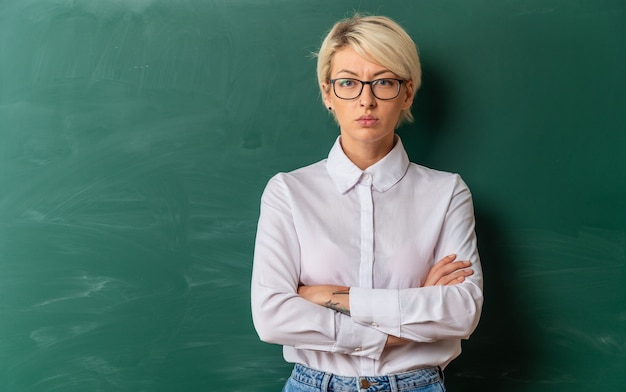 Jeune enseignante blonde stricte portant des lunettes en classe debout avec une posture fermée devant un tableau avec espace pour copie