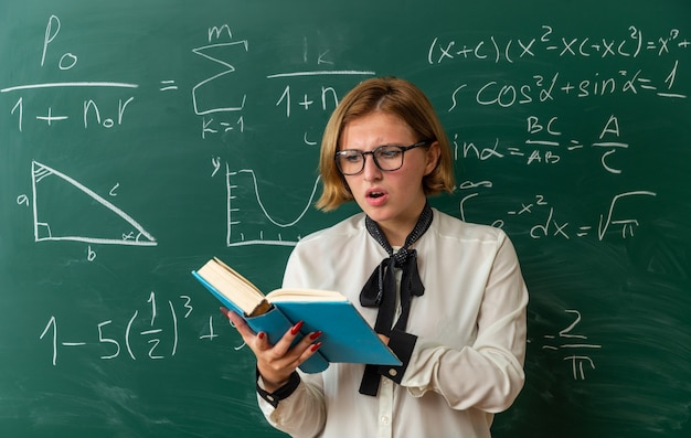 Jeune enseignante concernée portant des lunettes debout devant le livre de lecture du tableau noir en classe