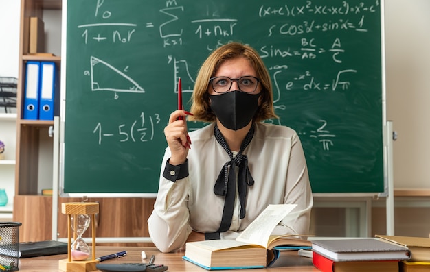 Une jeune enseignante concernée portant des lunettes et un masque médical est assise à table avec des outils scolaires tenant un crayon en classe