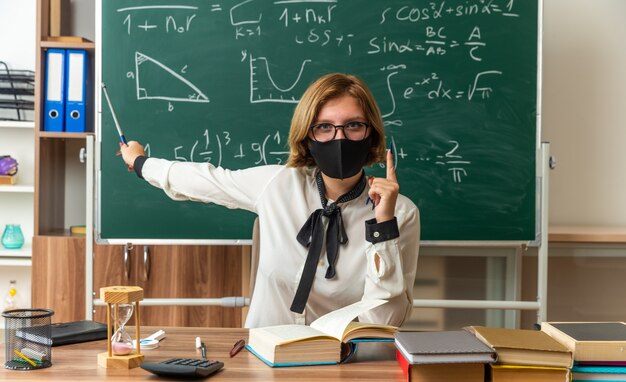 Une jeune enseignante confiante portant des lunettes et un masque médical est assise à table avec des outils scolaires pointe sur un tableau noir avec un bâton de pointeur en classe