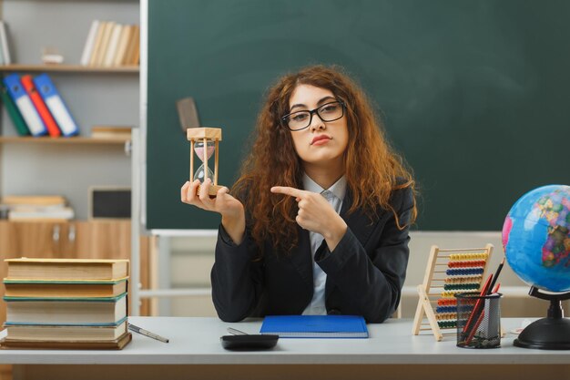 jeune enseignante confiante portant des lunettes tenant et pointant sur la montre de sable assise au bureau avec des outils scolaires en classe