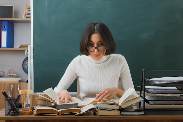 jeune enseignante confuse portant des lunettes lisant un livre assis au bureau avec des outils scolaires en classe