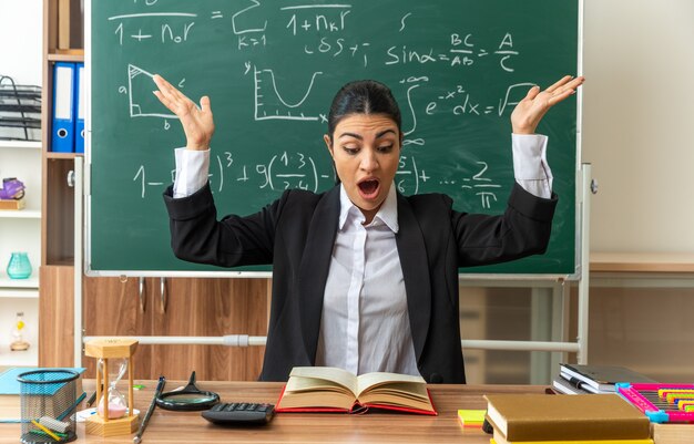 jeune enseignante effrayée est assise à table avec des fournitures scolaires livre de lecture écartant les mains en classe