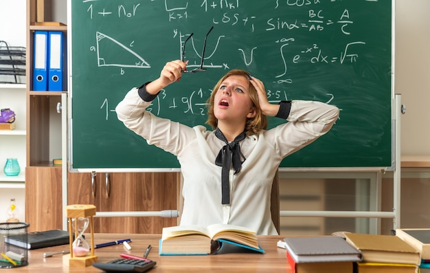 Une jeune enseignante effrayée est assise à table avec des fournitures scolaires tenant et regardant des lunettes mettant la main sur la tête en classe