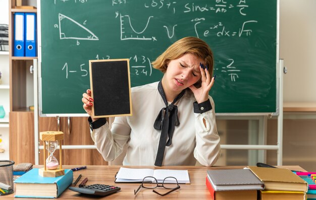 jeune enseignante est assise à table avec des fournitures scolaires tenant un mini tableau noir en classe