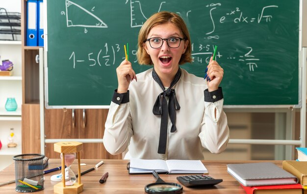 Jeune enseignante excitée est assise à table avec des fournitures scolaires tenant un crayon en classe