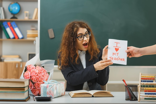 une jeune enseignante excitée a reçu une carte postale assise au bureau avec des outils scolaires en classe