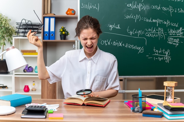 Jeune Enseignante De Mathématiques Agacée Enlevant Des Lunettes Assise Au Bureau Avec Des Fournitures Scolaires Tenant Une Loupe Gardant La Main Sur Un Livre Ouvert Criant Les Yeux Fermés Dans La Salle De Classe