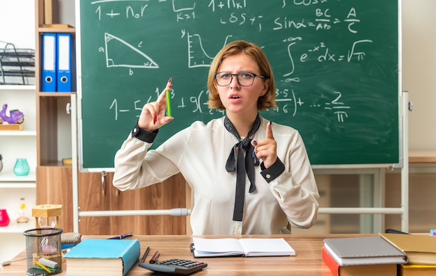 Une jeune enseignante mécontente est assise à table avec des fournitures scolaires tenant un crayon vous montrant un geste en classe