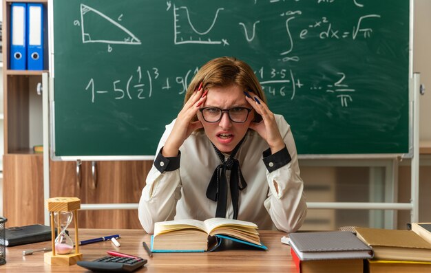 Une jeune enseignante mécontente portant des lunettes est assise à table avec des outils scolaires mettant les mains sur les temples en classe