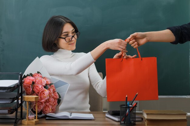 une jeune enseignante souriante a reçu un sac cadeau assis au bureau avec des outils scolaires en classe