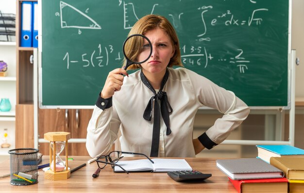 Une jeune enseignante stricte est assise à table avec des fournitures scolaires regardant à l'avant avec une loupe mettant la main sur la hanche en classe