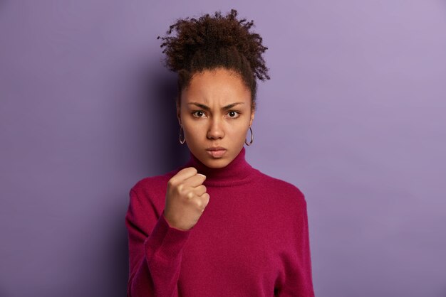 Une jeune enseignante stricte montre le poing fermé, le mécontentement du comportement des élèves, promet de punir, regarde en colère, a les cheveux bouclés, porte un pull bordeaux, isolé sur un mur violet