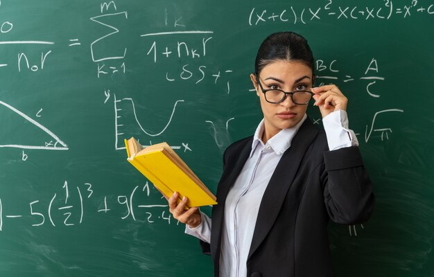 Jeune enseignante stricte portant des lunettes debout devant le tableau noir tenant un livre en classe