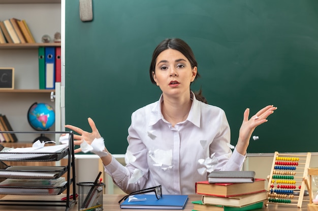 Jeune enseignante surprise écrasant du papier assis à table avec des outils scolaires en classe