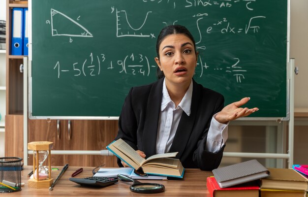 une jeune enseignante surprise est assise à table avec des fournitures scolaires écartant la main dans la salle de classe