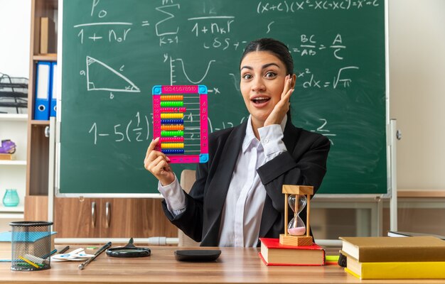 une jeune enseignante surprise est assise à table avec des fournitures scolaires tenant un boulier mettant la main sur la joue en classe