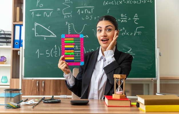 une jeune enseignante surprise est assise à table avec des fournitures scolaires tenant un boulier mettant la main sur la joue en classe