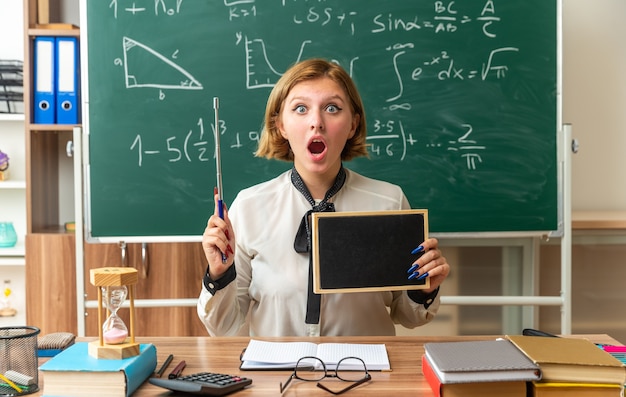 Photo gratuite une jeune enseignante surprise est assise à table avec des fournitures scolaires tenant un mini tableau noir avec un bâton de pointeur en classe