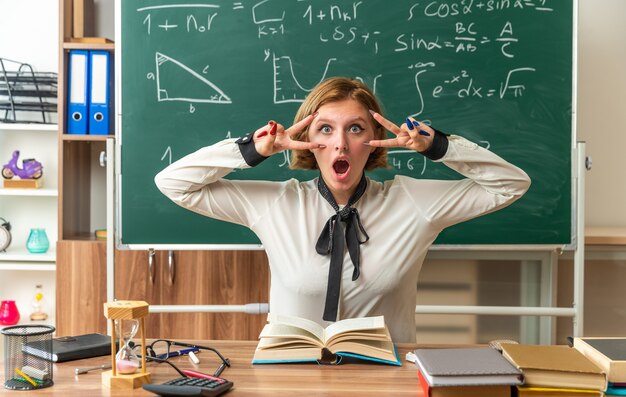 Une jeune enseignante surprise est assise à table avec des outils scolaires montrant un geste de paix en classe