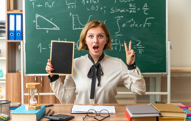 Une jeune enseignante surprise est assise à table avec des outils scolaires tenant un mini tableau noir montrant deux en classe