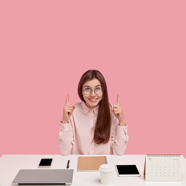 Jeune entrepreneur prospère prospère avec un regard heureux, pointe les deux index vers le plafond, porte une chemise élégante et des lunettes