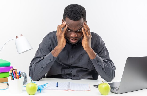 Jeune étudiant afro-américain agacé assis au bureau avec des outils scolaires mettant les mains sur son front