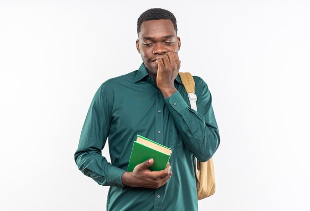 Jeune étudiant afro-américain anxieux avec sac à dos tenant un livre et se mordant les doigts