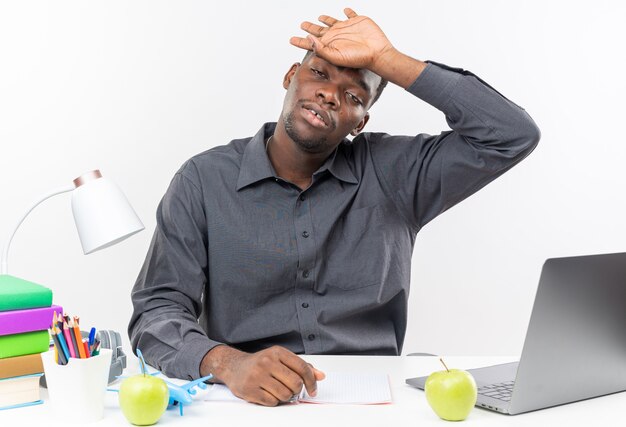 Jeune étudiant afro-américain fatigué assis au bureau avec des outils scolaires mettant la main sur son front isolé sur un mur blanc