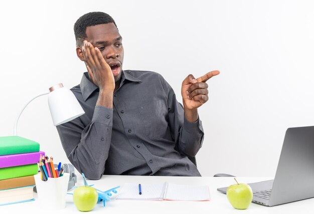 Jeune étudiant afro-américain surpris assis au bureau avec des outils scolaires mettant la main sur son visage regardant et pointant sur le côté