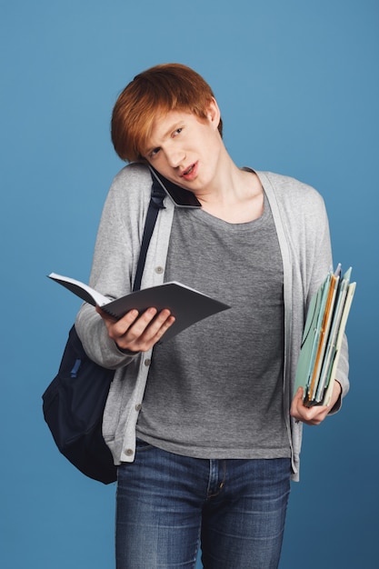 Jeune étudiant aux cheveux roux beau en tenue décontractée avec sac à dos noir tenant beaucoup de livres et cahier dans les mains, parler au téléphone avec la mère.