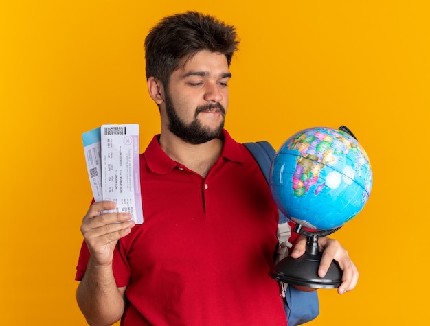 Jeune étudiant barbu guy en polo rouge avec sac à dos tenant globe et billets d'avion à la recherche intriguée et heureux debout sur fond orange