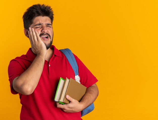 Jeune étudiant barbu guy en polo rouge avec sac à dos tenant des livres à la recherche de mal à toucher sa joue mal de dents debout sur fond orange