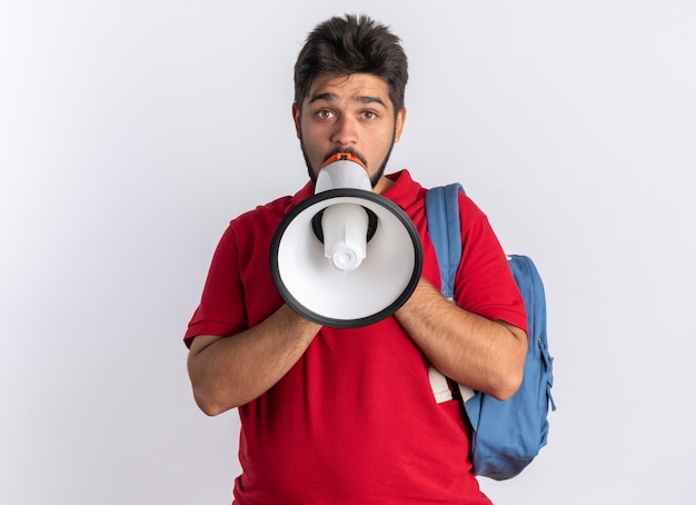 Jeune étudiant barbu en polo rouge avec sac à dos criant au mégaphone étant émotif et inquiet debout