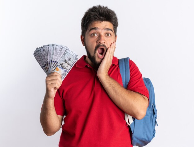 Jeune étudiant barbu en polo rouge avec sac à dos tenant de l'argent à l'air étonné et surpris debout