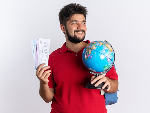 Jeune étudiant barbu en polo rouge avec sac à dos tenant des billets d'avion et globe regardant de côté avec le sourire sur un visage heureux debout sur un mur blanc