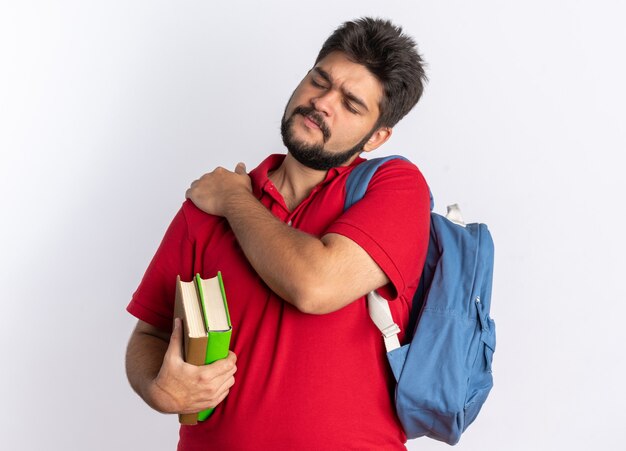 Jeune étudiant barbu en polo rouge avec sac à dos tenant des cahiers ayant l'air malade de toucher son épaule ressentant une douleur debout