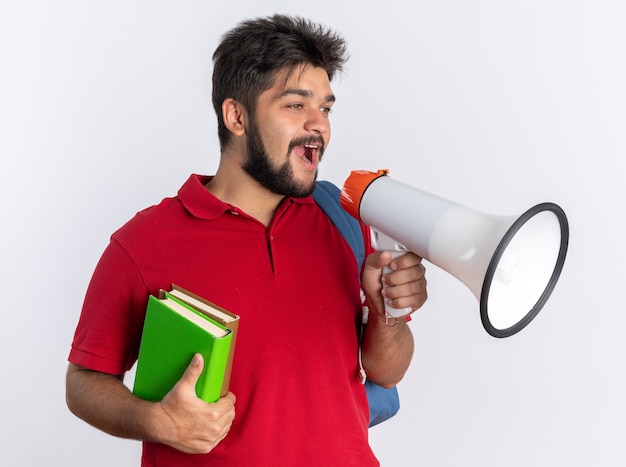 Jeune étudiant barbu en polo rouge avec sac à dos tenant des cahiers criant au mégaphone debout heureux et positif