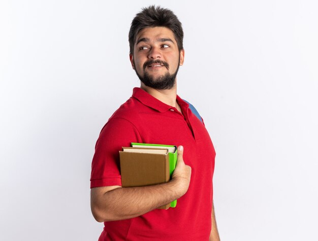 Jeune étudiant barbu en polo rouge avec sac à dos tenant des cahiers regardant de côté souriant confiant debout sur un mur blanc