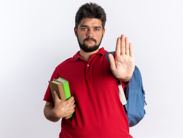 Jeune étudiant barbu en polo rouge avec sac à dos tenant des cahiers avec un visage sérieux faisant un geste d'arrêt avec la main debout sur un mur blanc