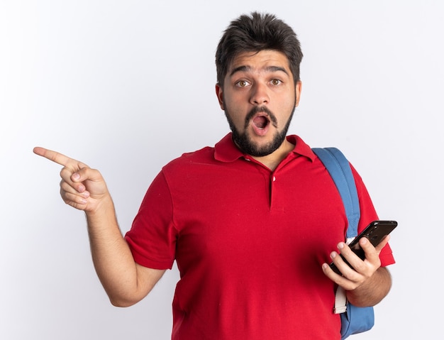 Jeune étudiant barbu en polo rouge avec sac à dos tenant un smartphone à la surprise de pointer avec l'index sur le côté debout sur un mur blanc