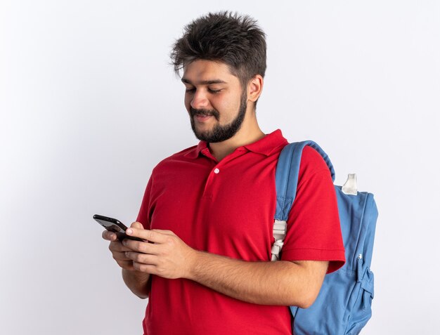 Jeune étudiant barbu en polo rouge avec sac à dos tenant un smartphone en tapant des messages souriant confiant debout sur un mur blanc