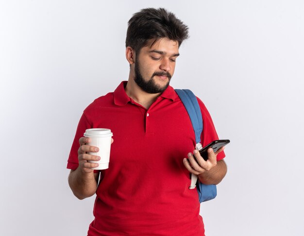 Jeune étudiant barbu en polo rouge avec sac à dos tenant un smartphone et une tasse de café à l'air confiant souriant debout sur un mur blanc