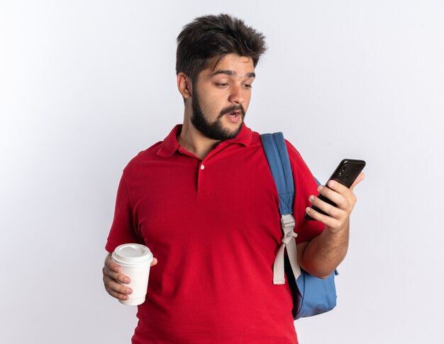 Jeune étudiant barbu en polo rouge avec sac à dos tenant un smartphone et une tasse de café à la confusion debout