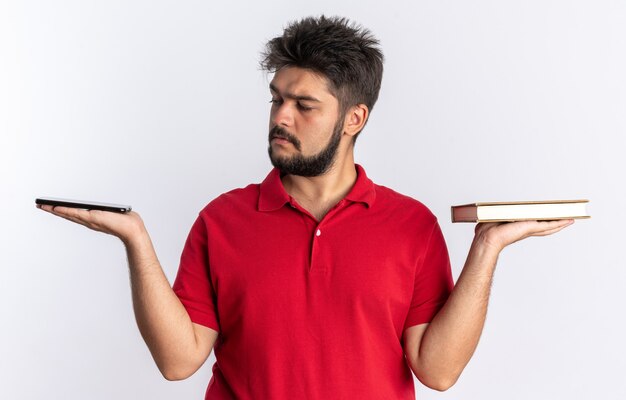 Jeune étudiant barbu en polo rouge tenant des cahiers dans les mains en les regardant avec un visage sérieux debout