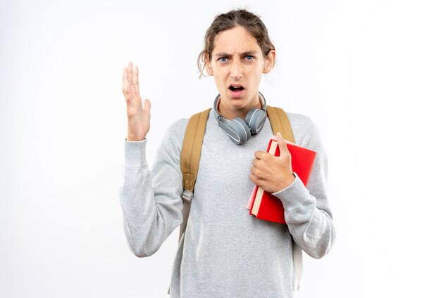 Jeune étudiant en colère portant un sac à dos avec un casque sur le cou tenant des livres isolés sur un mur blanc