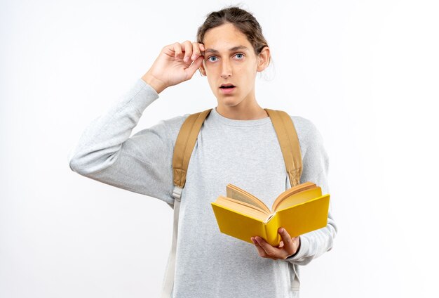 Jeune étudiant effrayé portant un sac à dos tenant un livre mettant la main sur le front isolé sur un mur blanc