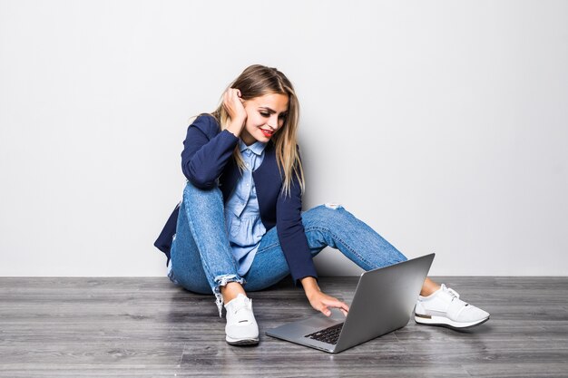 Jeune étudiant heureux assis sur le sol et en tapant sur un ordinateur portable sur fond gris